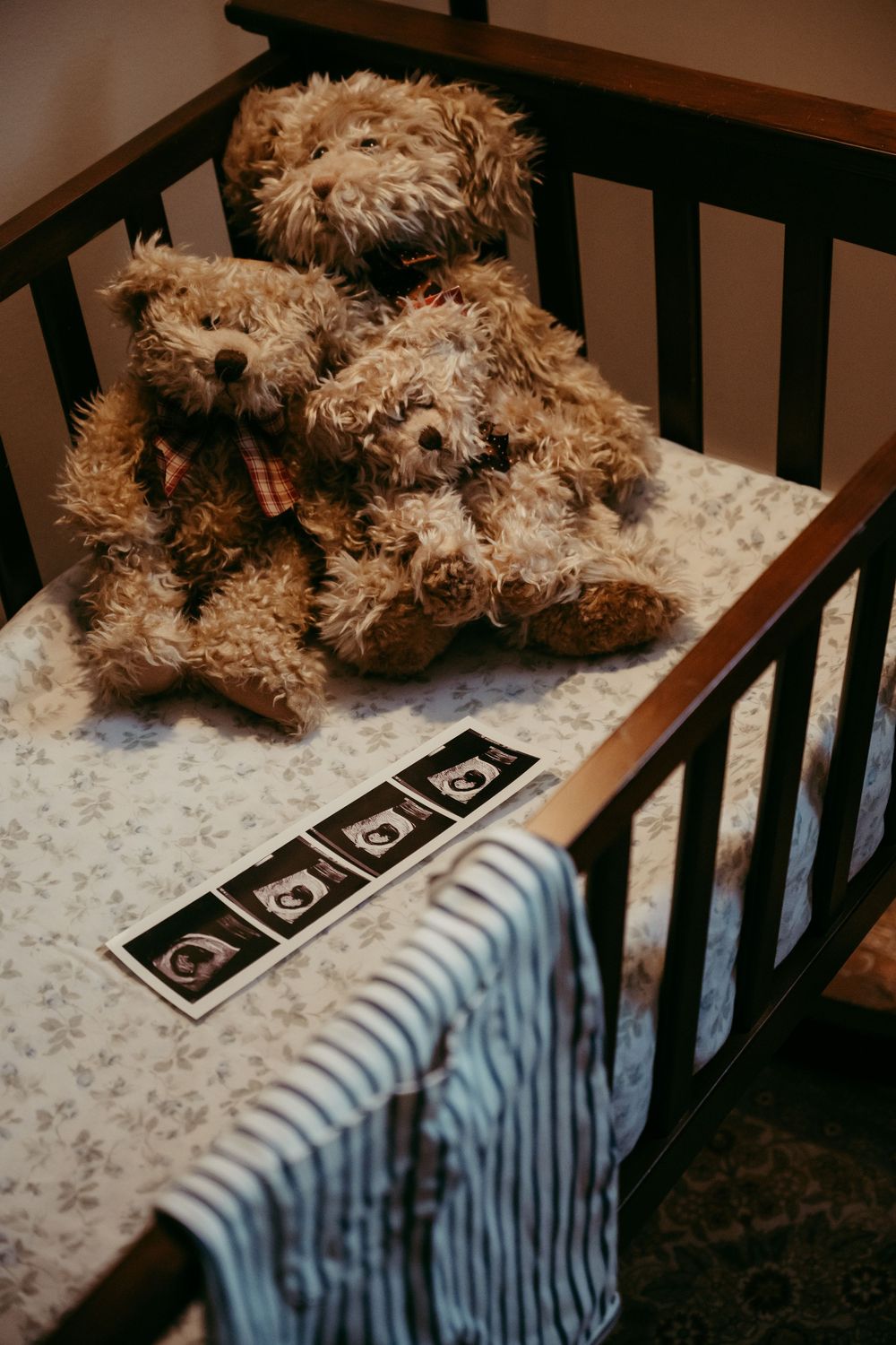 Teddy bears sitting in a baby crib with ultrasound photos and a striped blanket on floral bedding.
