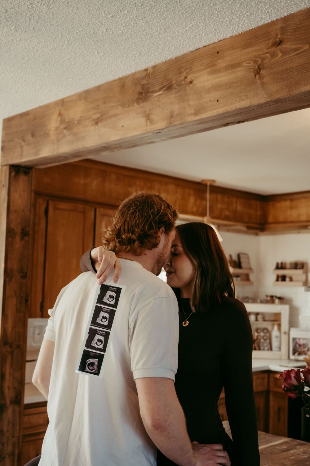 A couple shares a tender kiss in a rustic kitchen, the man wearing a white shirt with ultrasound photos on the back.