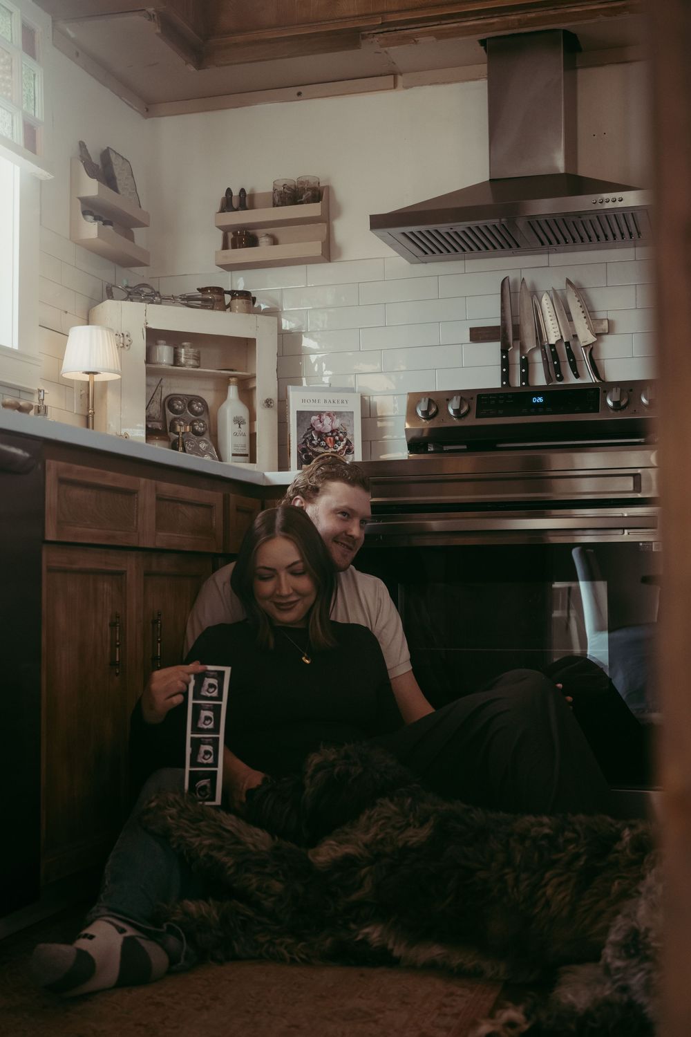 Couple sitting on kitchen floor with dog, holding ultrasound photos, announcing pregnancy in cozy home kitchen.