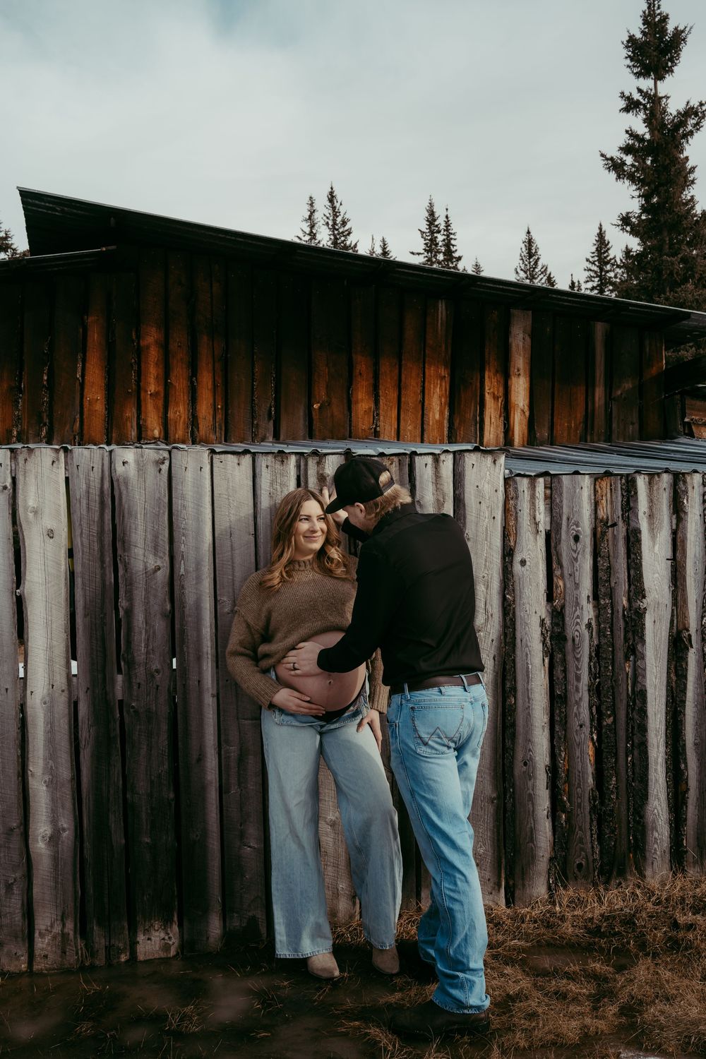Couple sharing a tender maternity moment against a rustic wooden fence with evergreen trees in the background.
