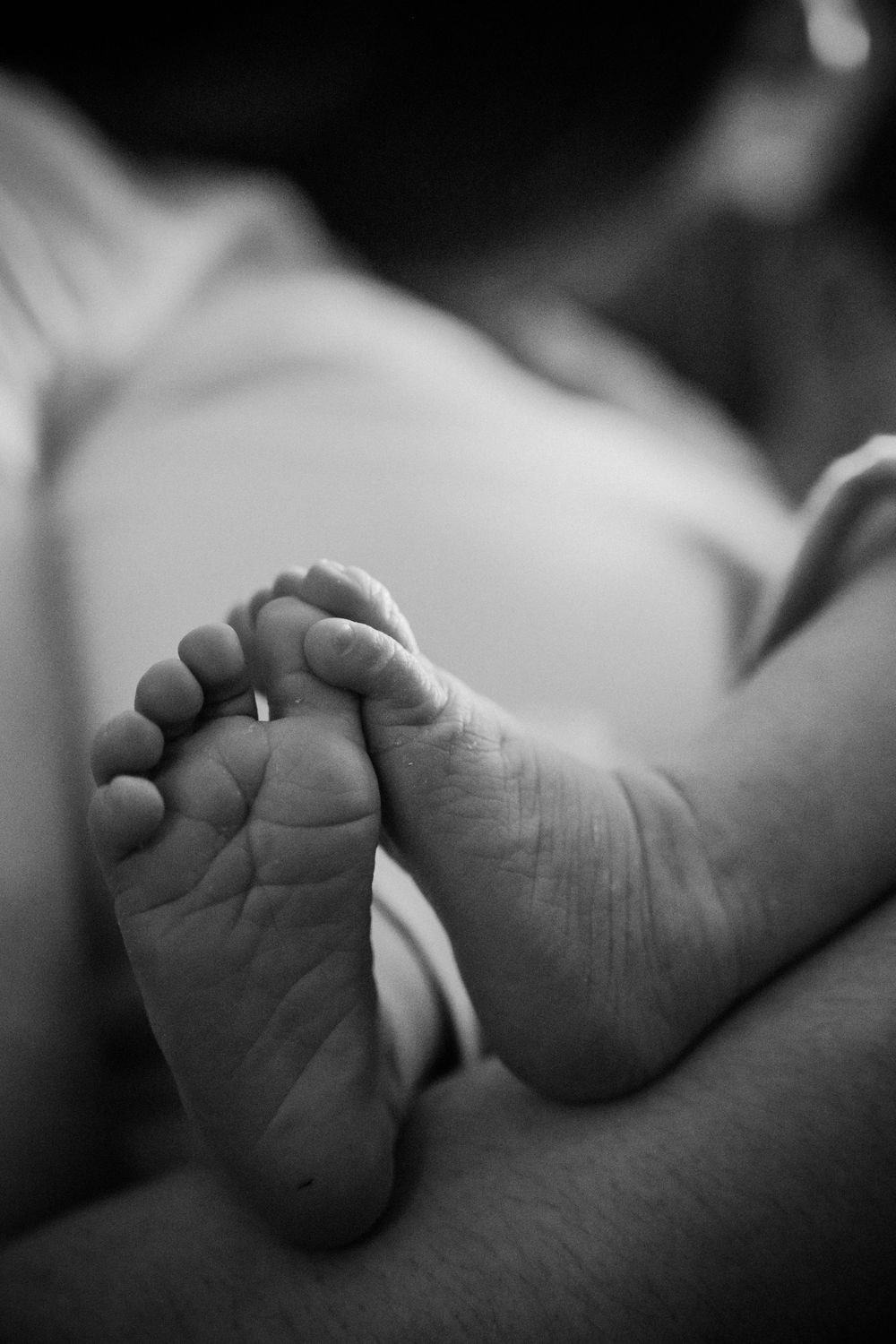 Close-up black and white photograph of a tiny baby's foot showing delicate toes and wrinkled sole against a soft background.