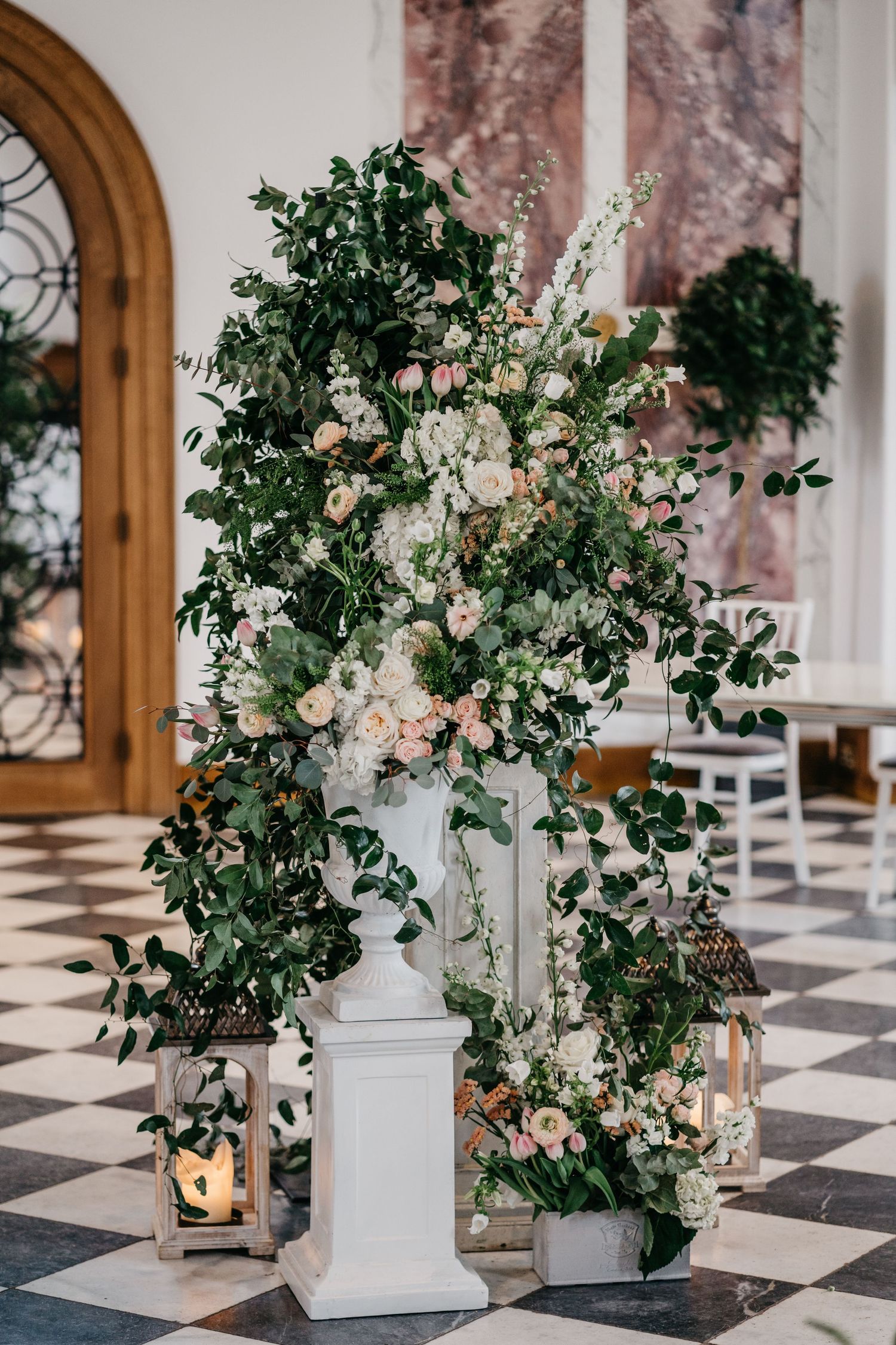 Elegant floral arrangement with greenery and roses on white pedestal with lantern decorations.