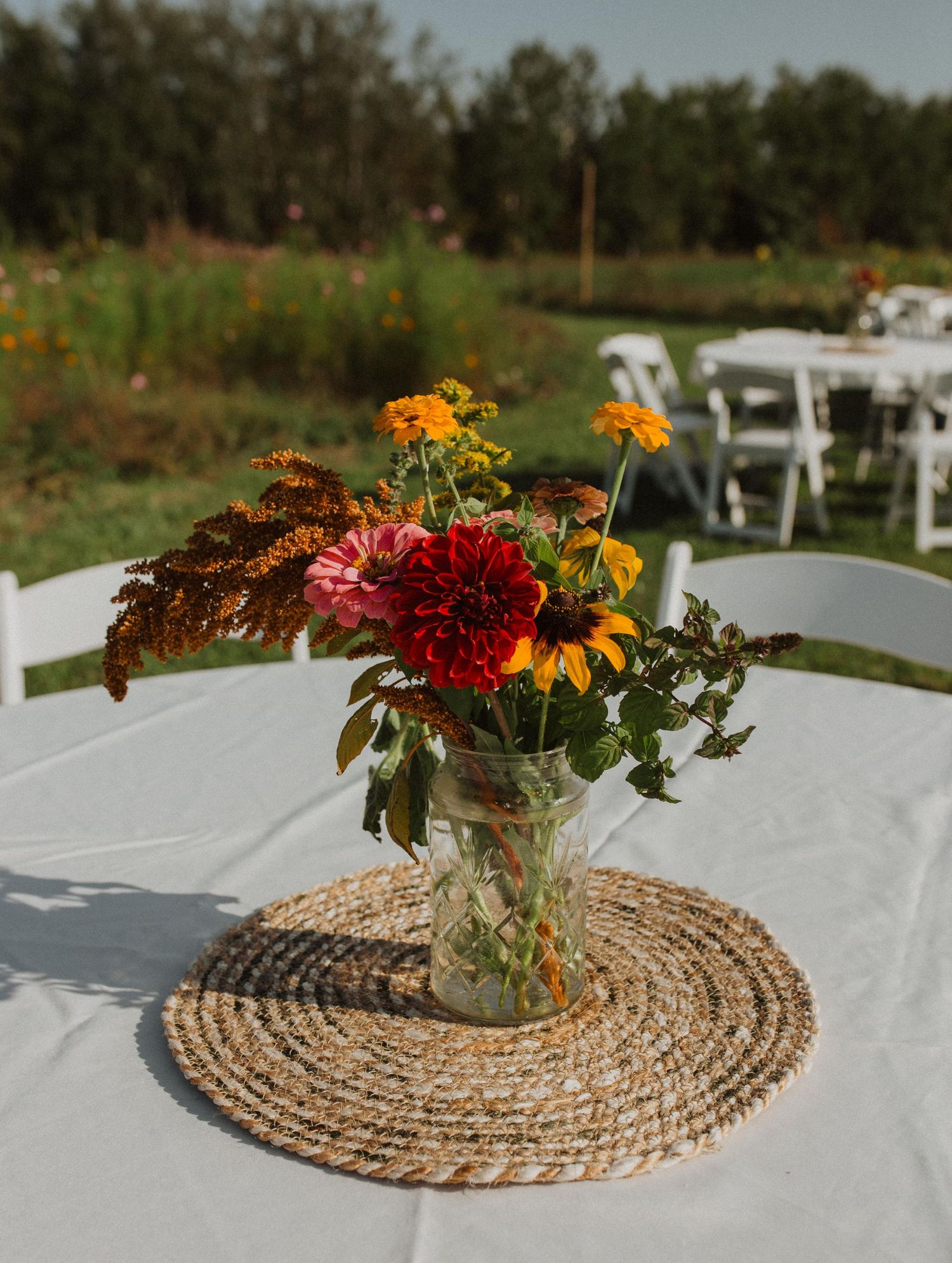 Mason jar centerpiece with red dahlias and sunflowers on a white tablecloth with woven placemat.