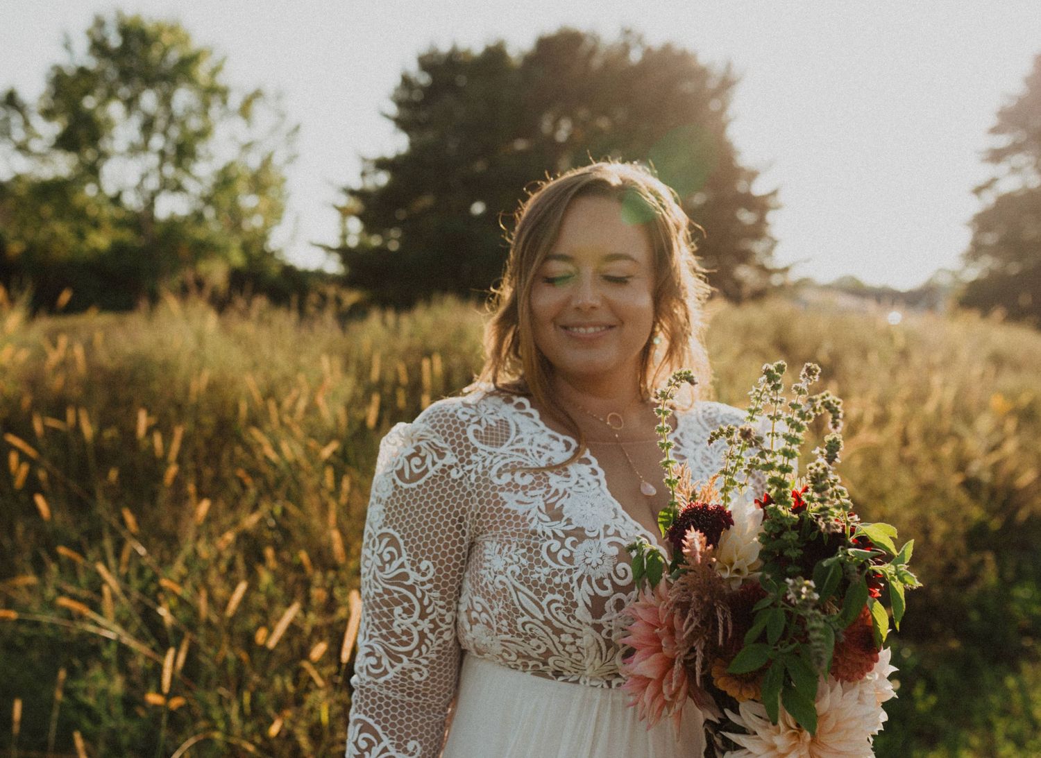 A bride in a lace wedding dress poses in a sunlit field during golden hour.