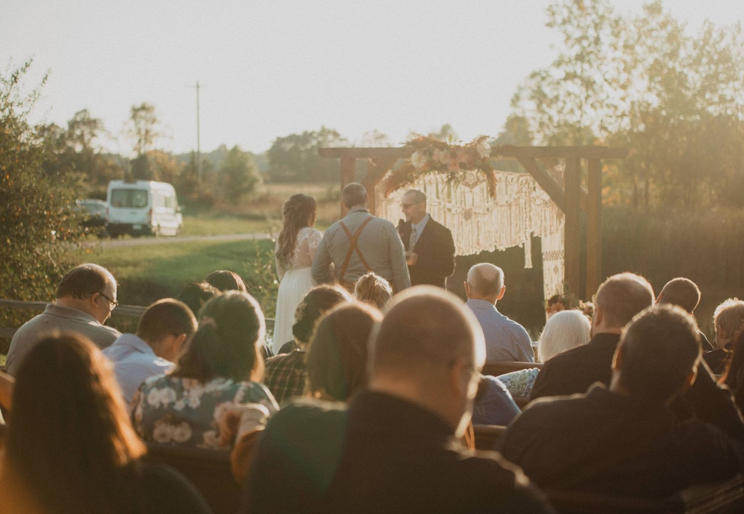 Wedding guests watch ceremony from wooden benches as golden sunlight illuminates outdoor lakeside venue.