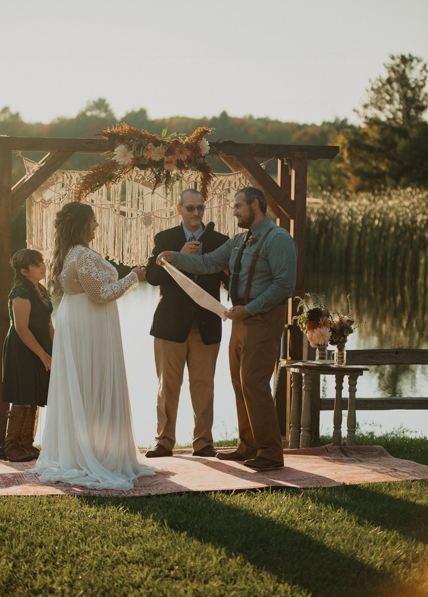 Outdoor wedding ceremony under macrame arch with autumn floral decorations by lakeside.