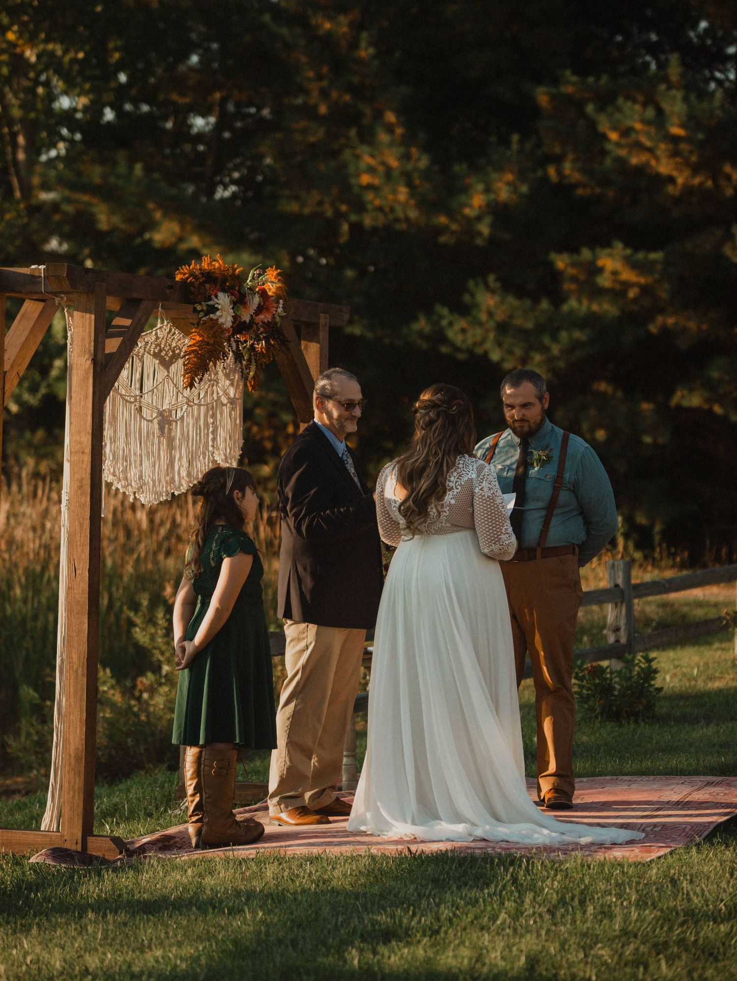 An intimate outdoor wedding ceremony takes place under a wooden arch decorated with macrame at golden hour.