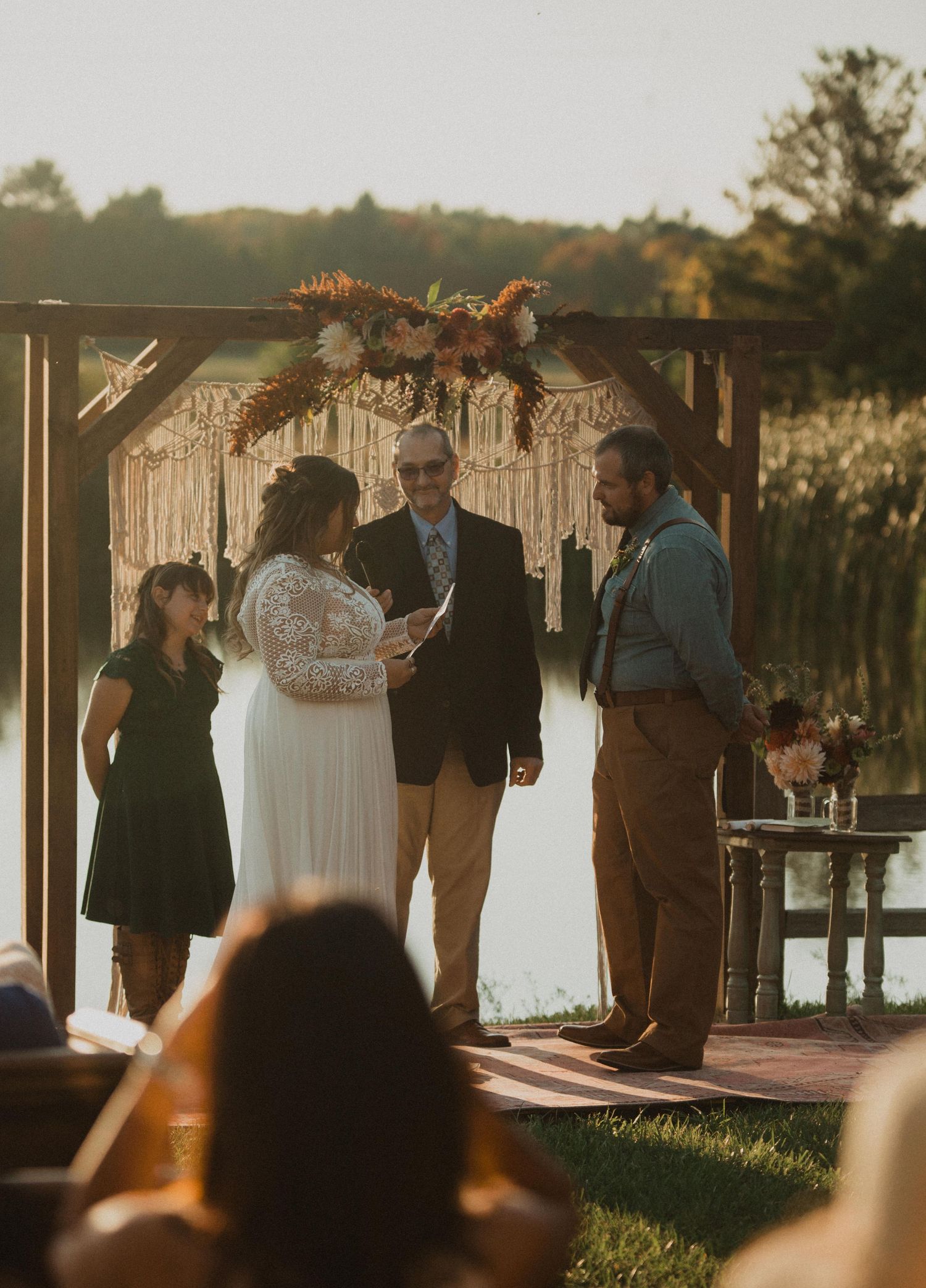A sunset wedding ceremony unfolds beneath a rustic wooden arch adorned with macrame and floral decorations.
