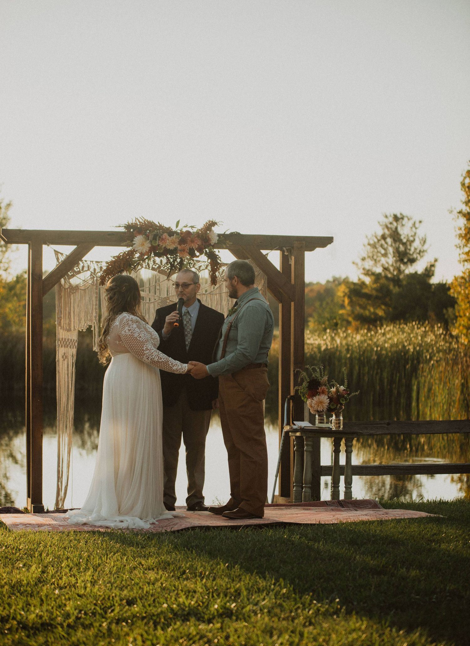 A wedding ceremony takes place by a lake at sunset under a wooden arch with macrame details.