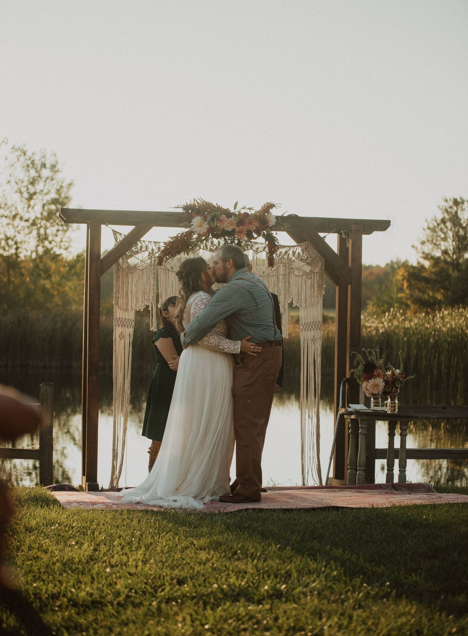 A tender moment during a lakeside wedding ceremony under a decorated wooden arch at sunset.
