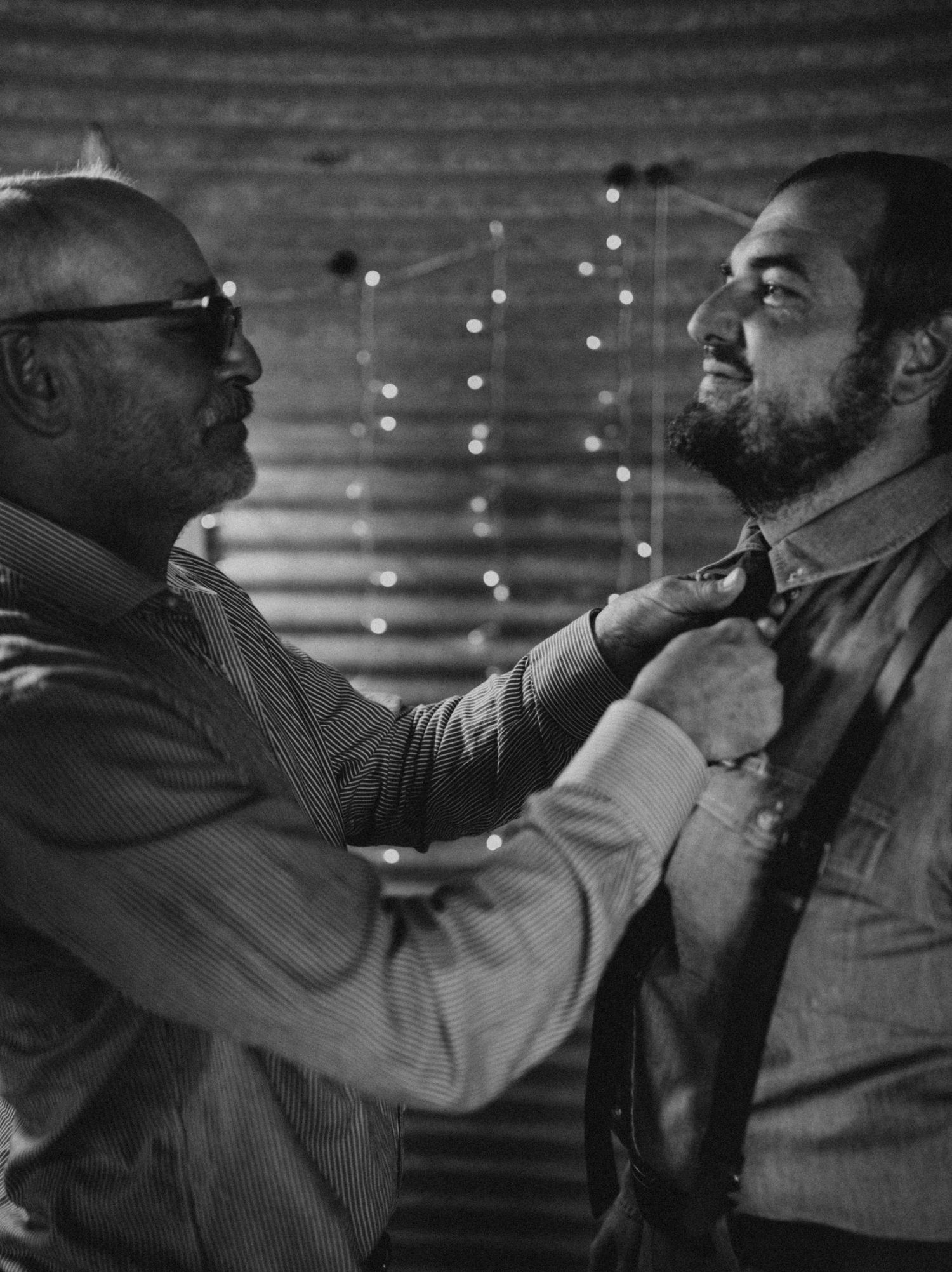 Two men share an emotional moment in front of a wall with twinkling lights in black and white.