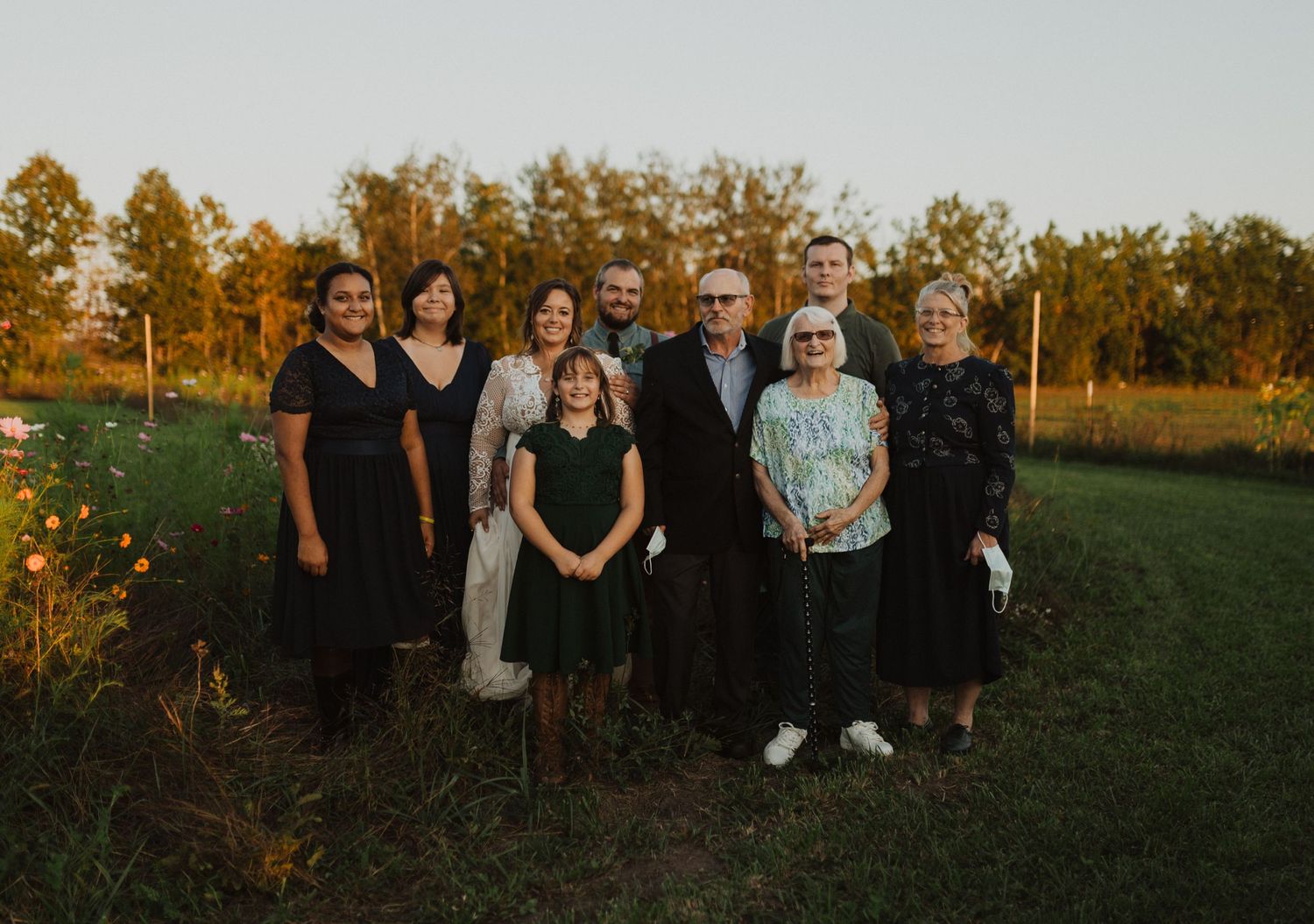Wedding group photo with attendees dressed in dark attire against sunset backdrop.