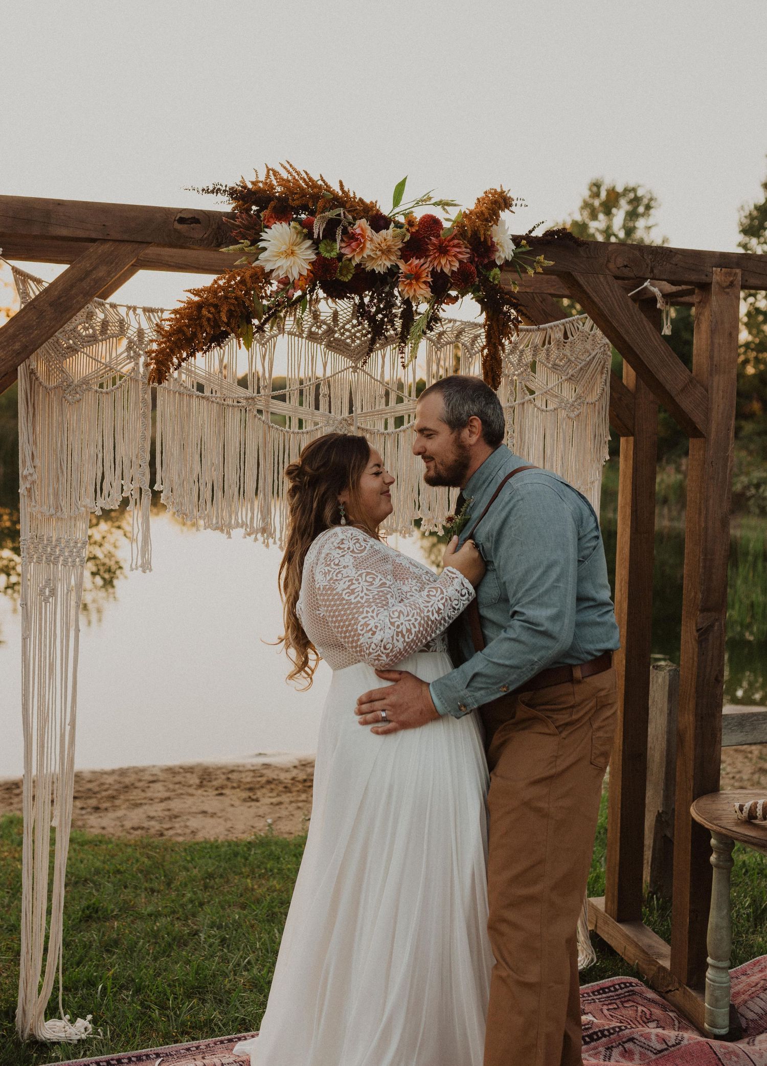 A wedding couple shares an intimate moment under a macrame arch decorated with autumn flowers at sunset.