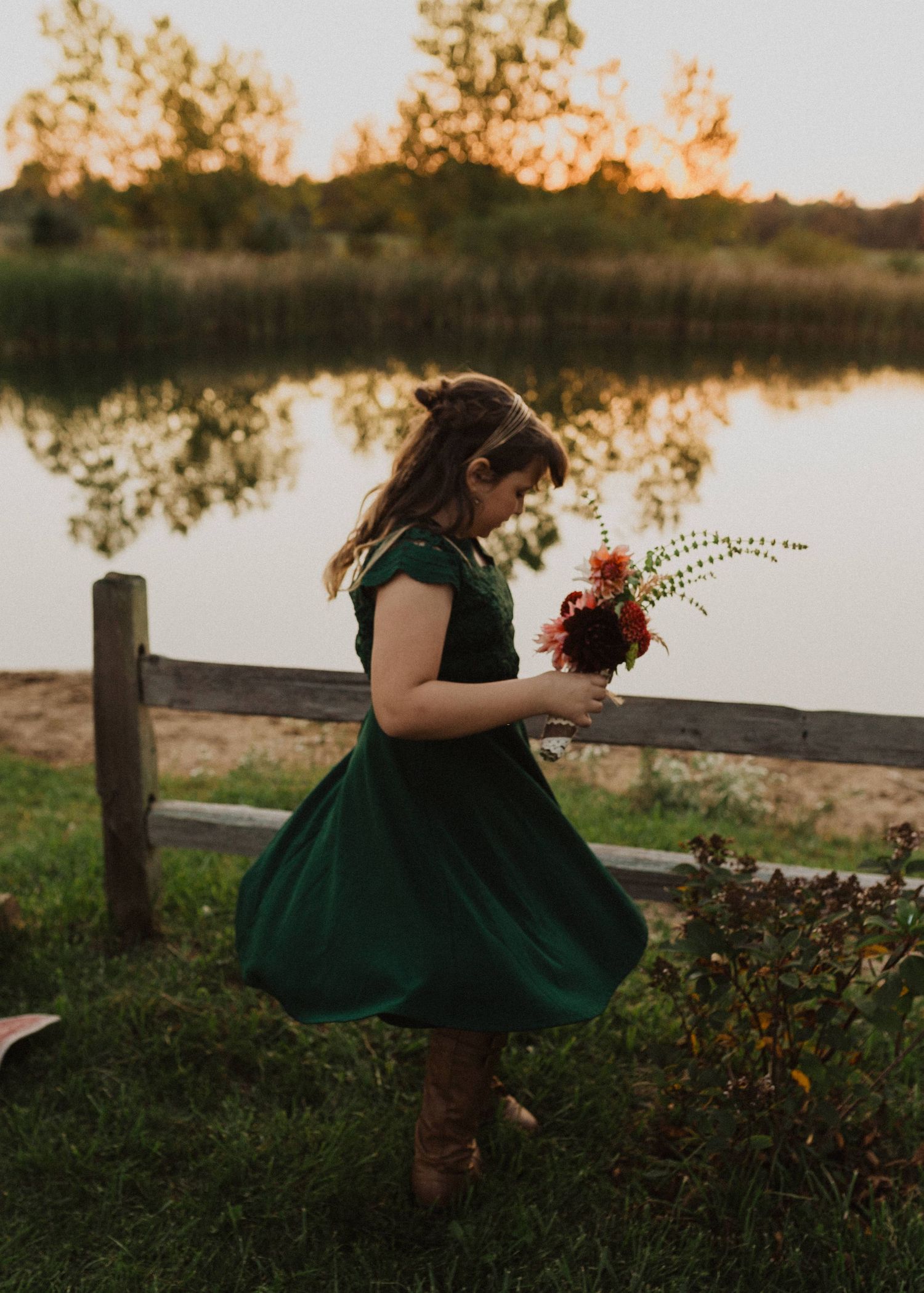A green dress contrasts against autumn grass near a lakeside wooden fence during sunset.