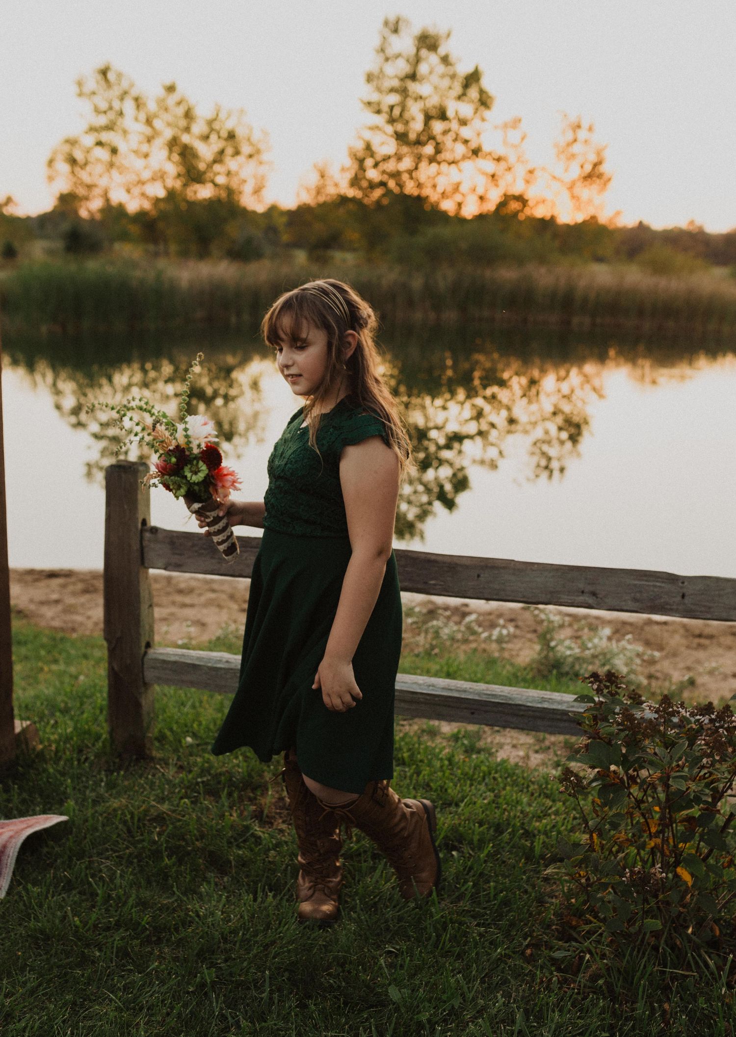 A person in emerald green stands near a rustic fence with a lake view at dusk.