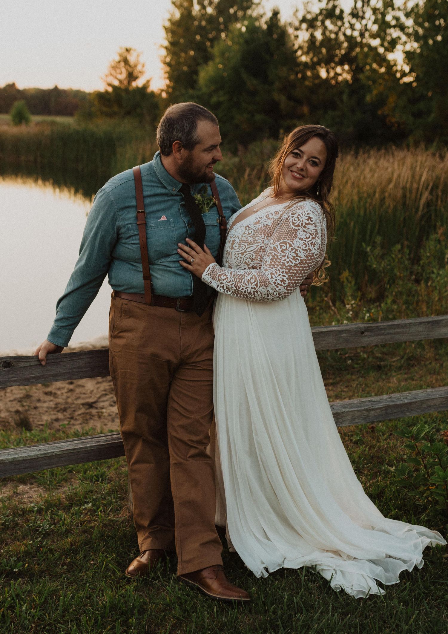 A joyful wedding couple laughs together by a lake during golden hour.