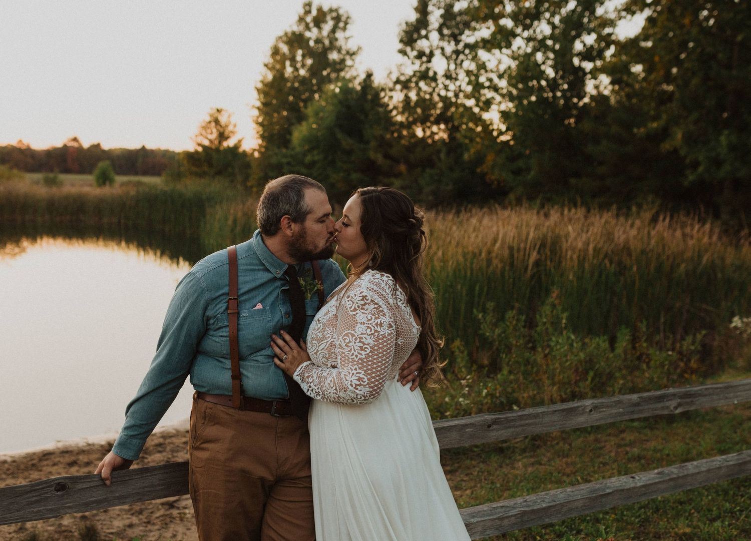 A romantic sunset photo of a couple by a lake with tall grass in the background.