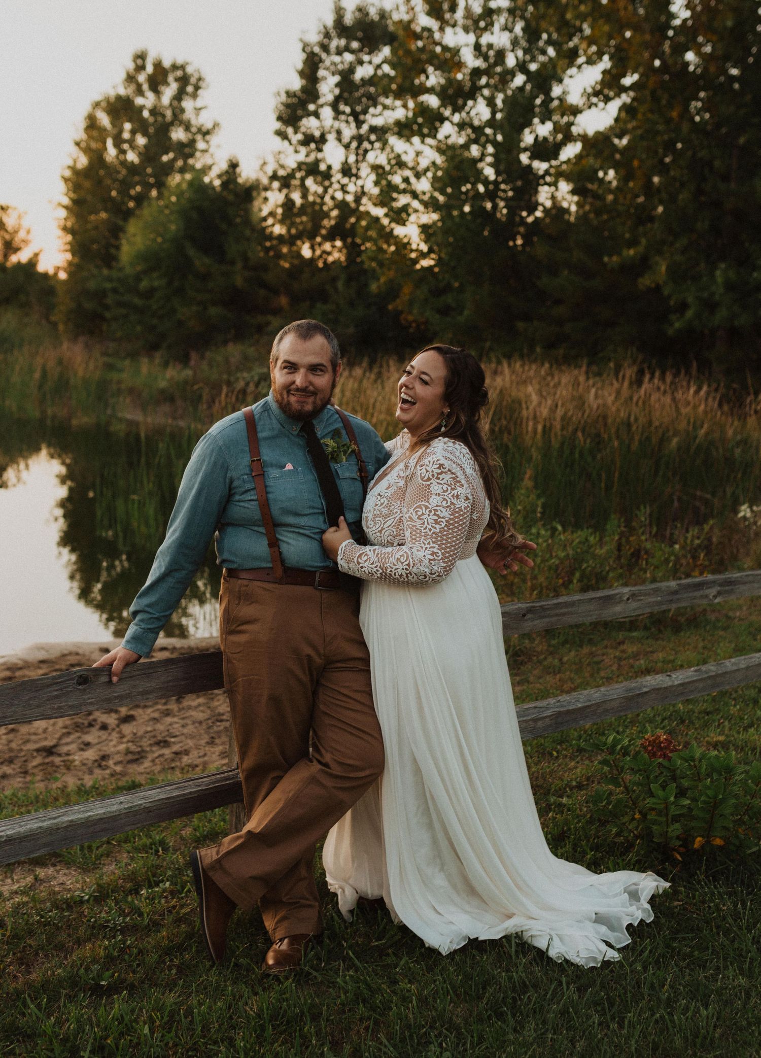 A couple shares a joyful moment at sunset by a lake, wearing a white lace dress and brown overalls.