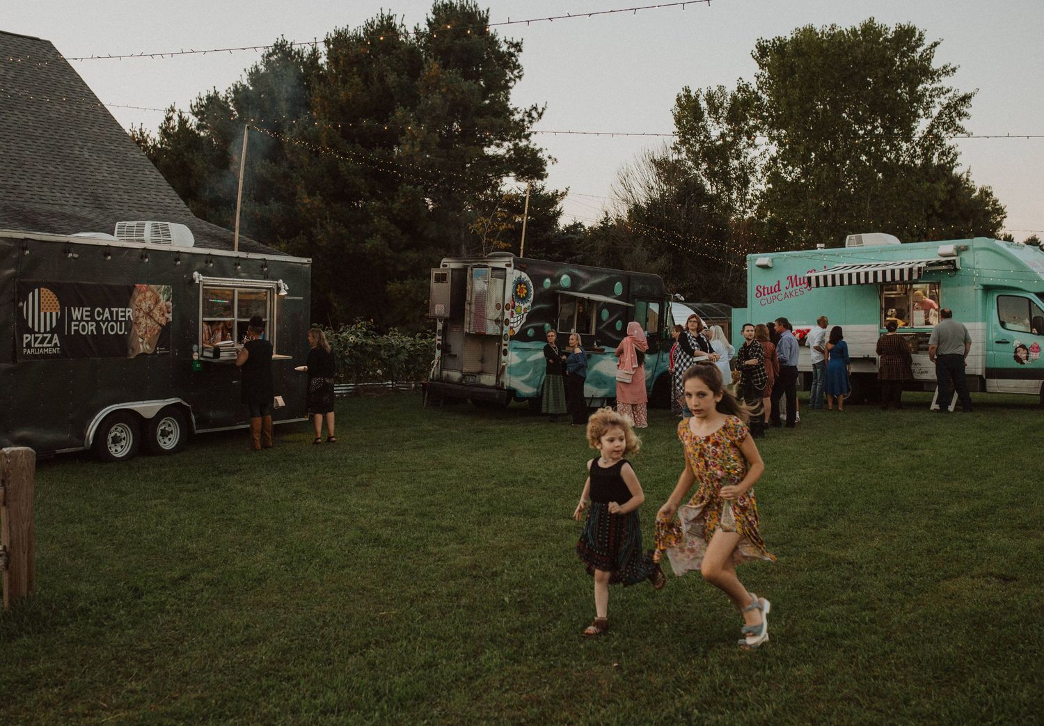Guests mingling at an outdoor event with food trucks and string lights at dusk.