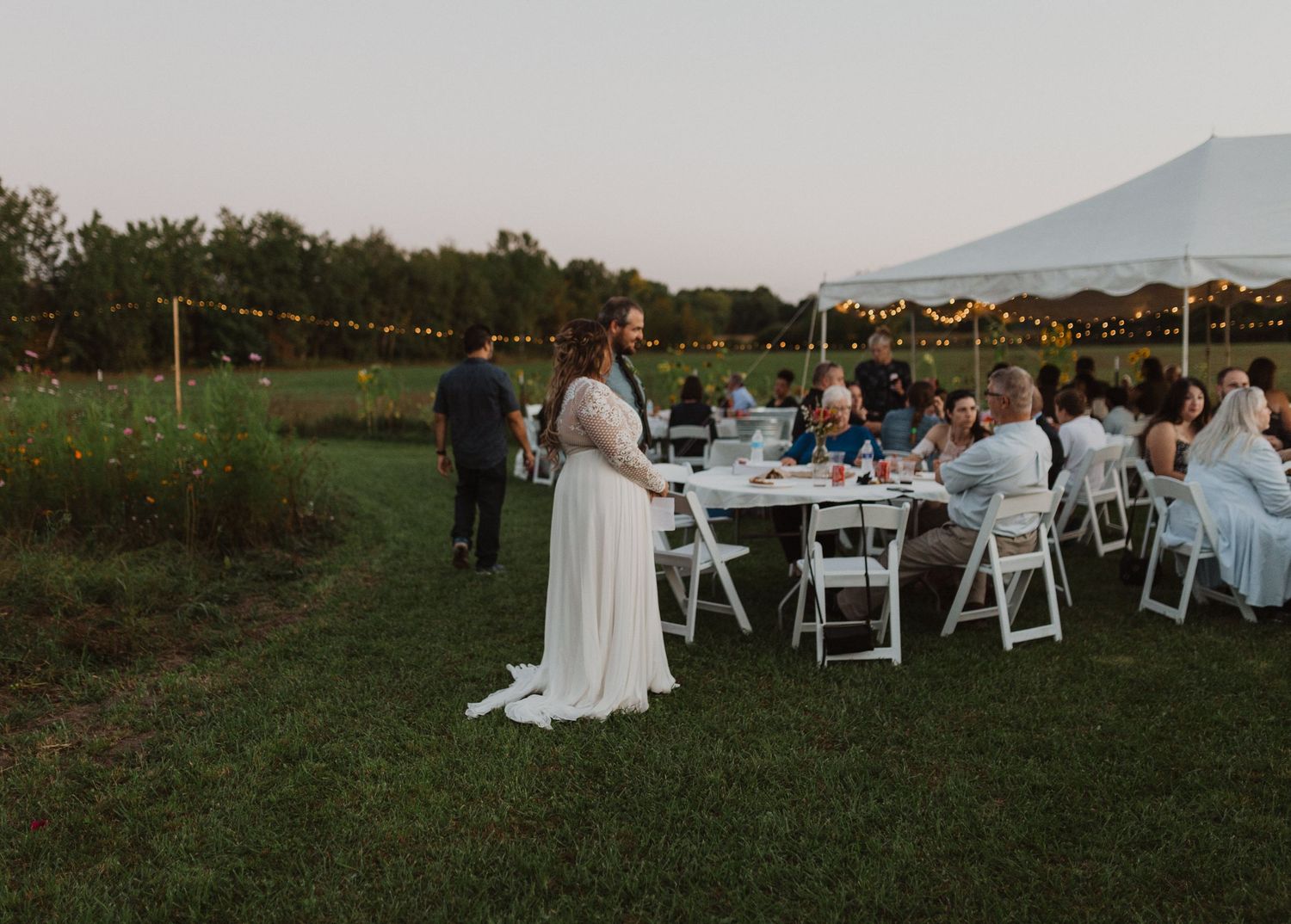 Outdoor wedding reception venue with white chairs and tent setup in a grassy field at dusk.