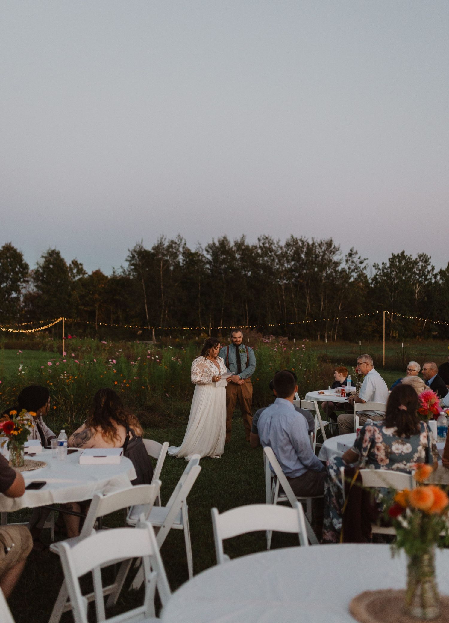 Wedding reception tables decorated with floral centerpieces and white chairs under string lights.