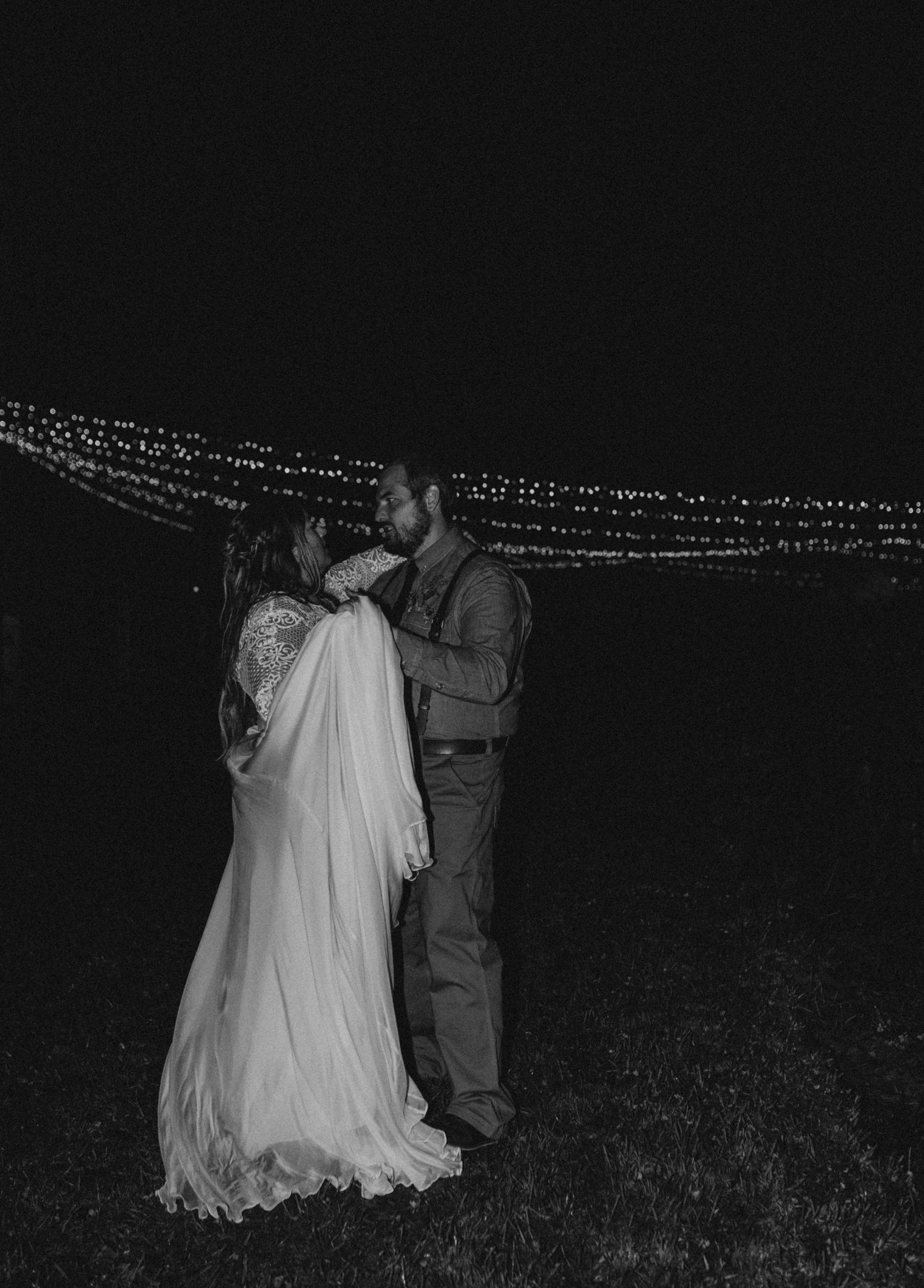 A black and white photo of a couple dancing outdoors under string lights at night.