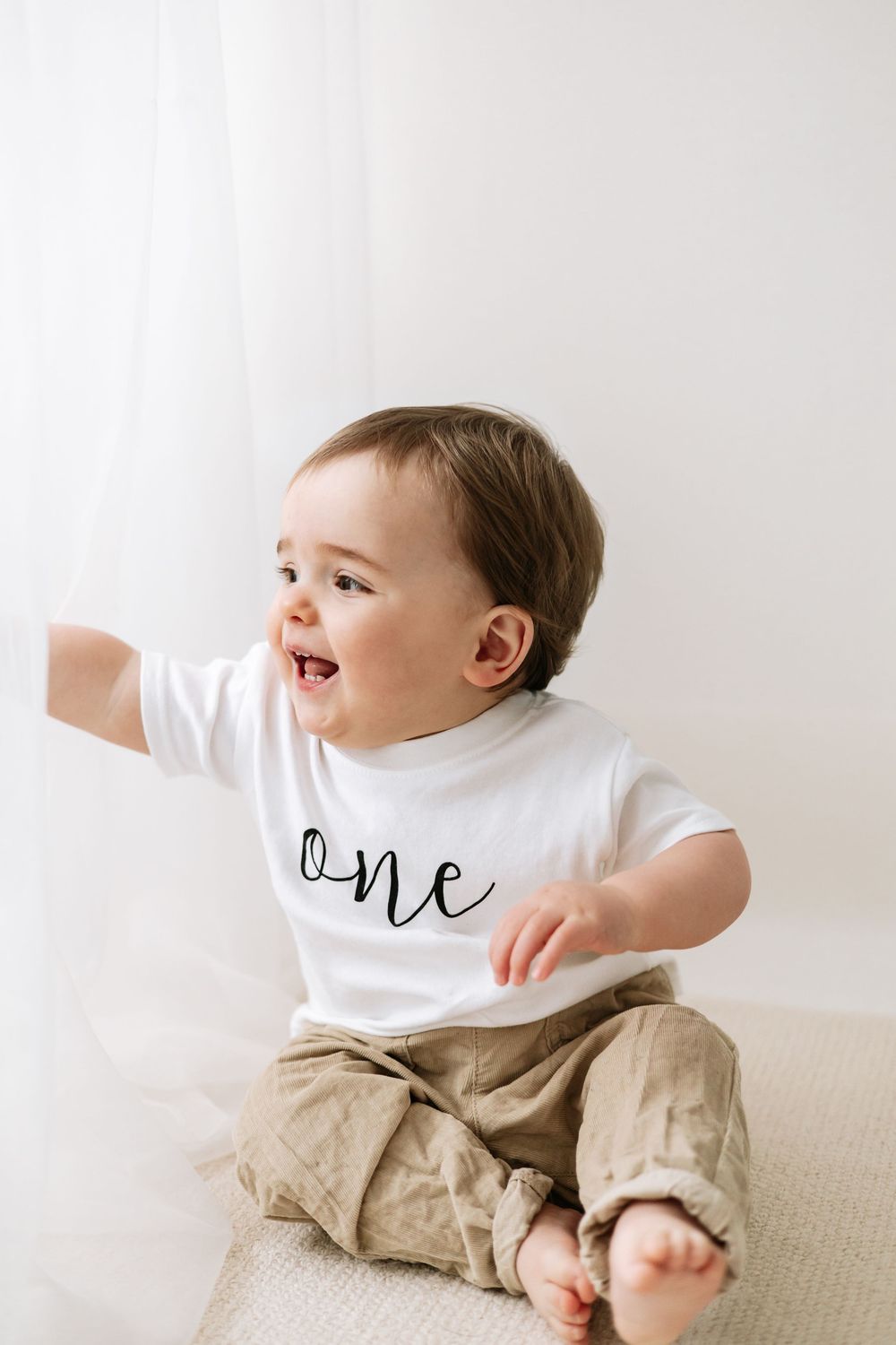 A happy baby wearing a white “one” shirt and sits smiling during a first birthday photo session Nottingham