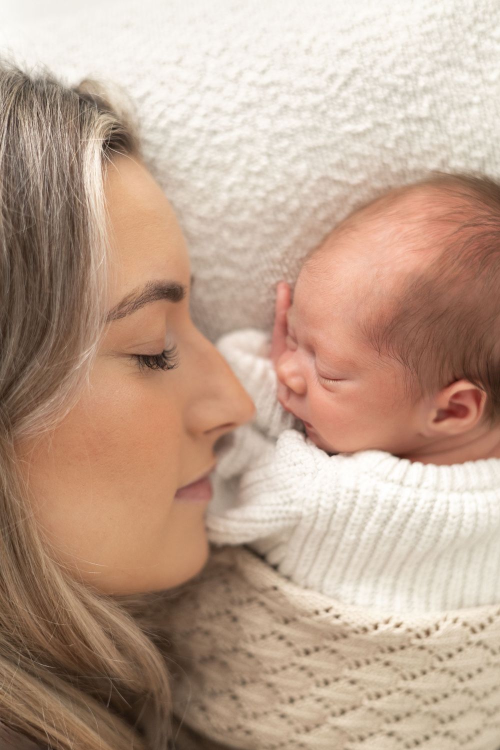 A mother gazes lovingly at her sleeping newborn baby wrapped in a white knit blanket natural newborn photography Nottingham NG16