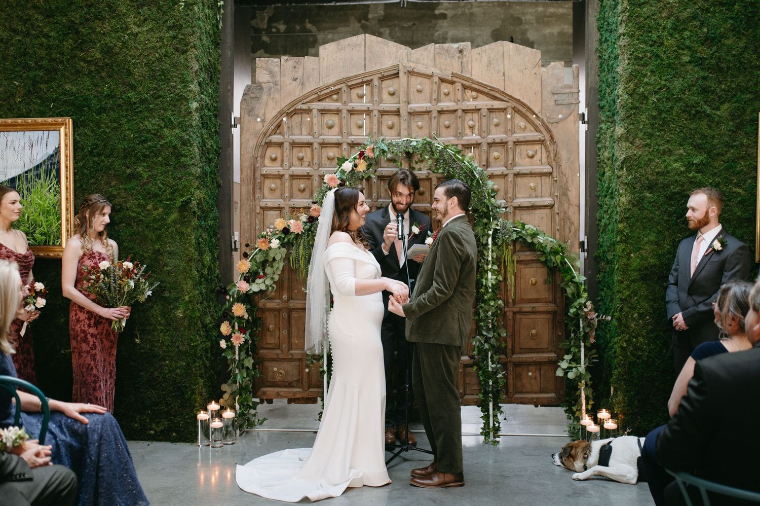 A wedding ceremony under an ornate wooden arch decorated with floral arrangements and greenery.