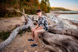 Senior boy in a floral shirt and jeans, sitting on driftwood on a beach at sunset, smiling confidently.