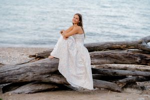 Smiling woman in a flowing white dress sits on a large driftwood log on a pebbly beach with calm water behind her.