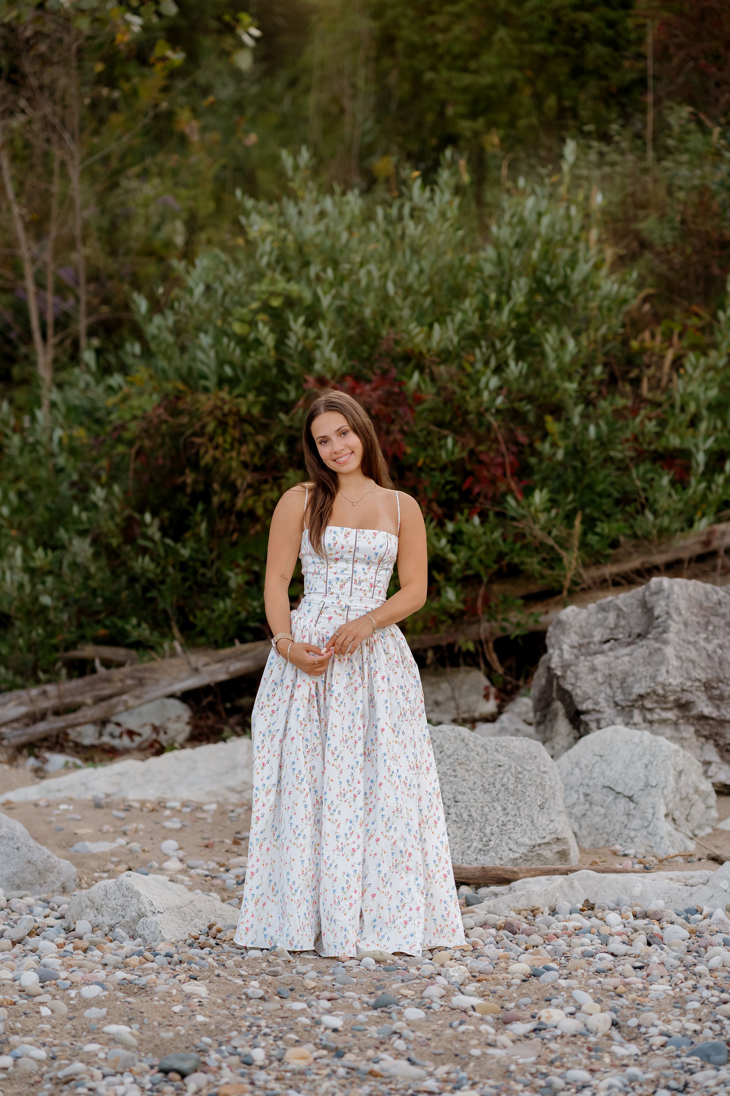 Smiling woman in a long white floral sundress stands on a rocky pebble beach with large boulders, driftwood and green shrubs behind her.