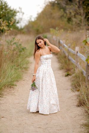 Young woman in a white floral sundress holding wildflowers while standing on a sandy path lined by a rustic fence and tall grasses.