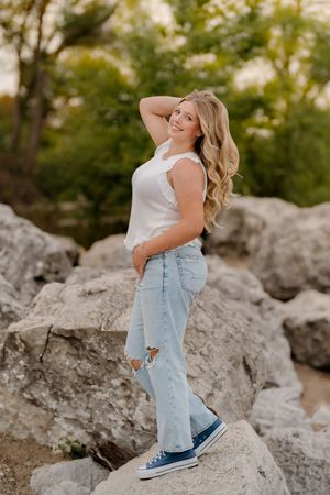 Smiling young woman with long wavy blonde hair poses on large rocks in a white top, ripped light-wash jeans and navy sneakers outdoors.