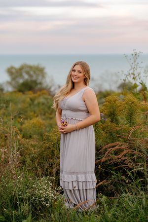 Smiling young woman with long blonde hair in a gray ruffled maxi dress holding wildflowers in a grassy field overlooking a lake at sunset.