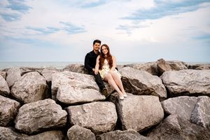 Two people sit close together on large rocky boulders along a coastline with cloudy sky in the background.