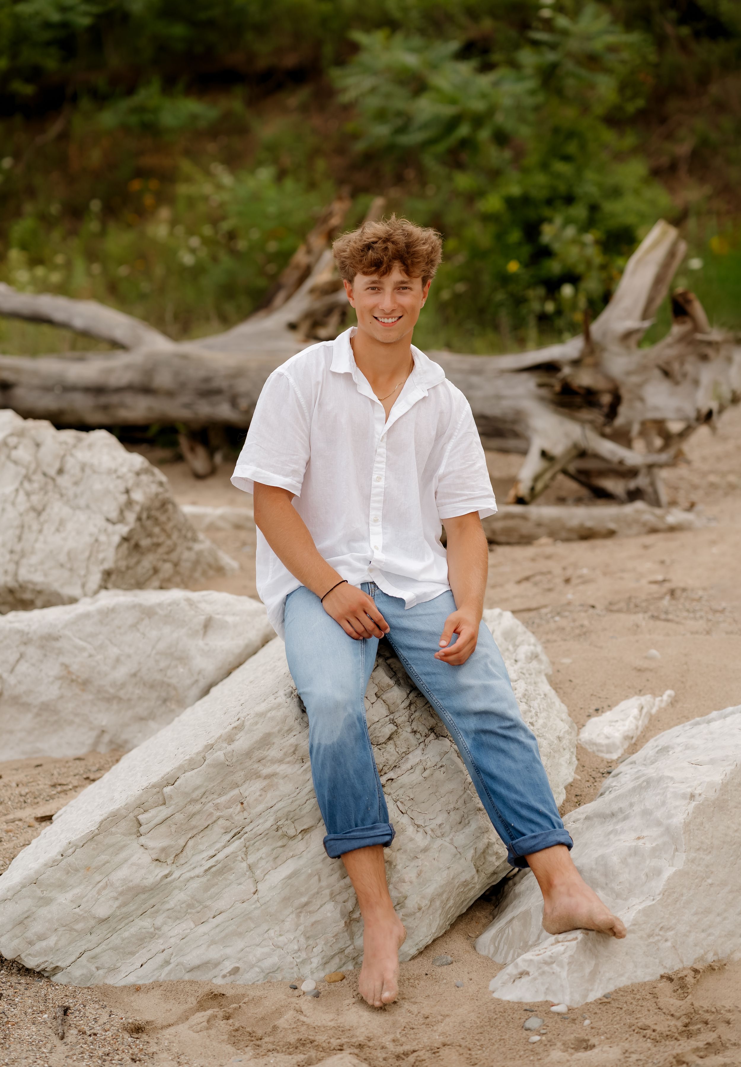 Smiling young man in white shirt and jeans sitting barefoot on rocks at a sandy beach with driftwood.
