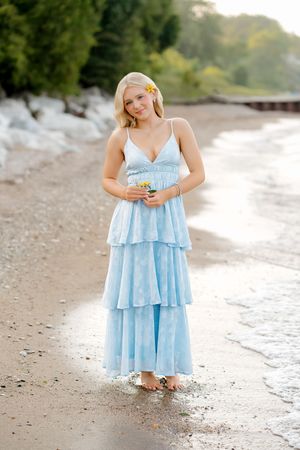 Blonde woman in blue tiered maxi dress with flower in hair standing barefoot on sandy beach shore.