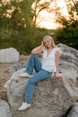 Young blonde woman sitting on rocks at golden hour, wearing white top and blue jeans, smiling outdoors.