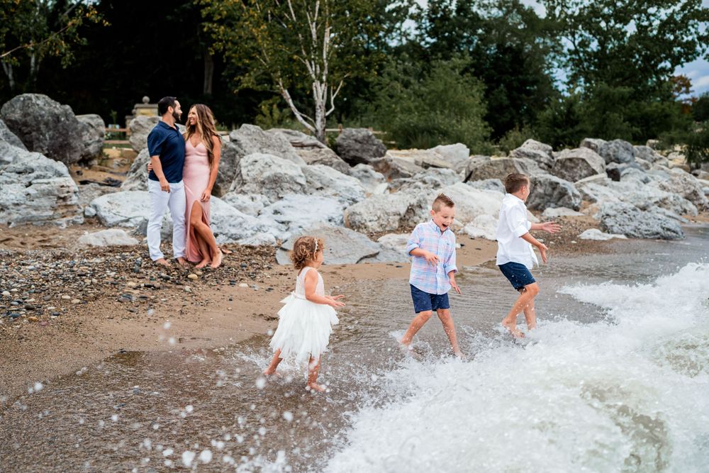 Children play in splashing waves while adults watch from rocky beach terrain.