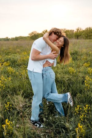 A couple embraces in a meadow of yellow wildflowers during golden hour while wearing casual denim and white outfits.