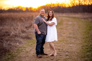 A couple poses together on a dirt path at sunset with golden light glowing through trees in the background.
