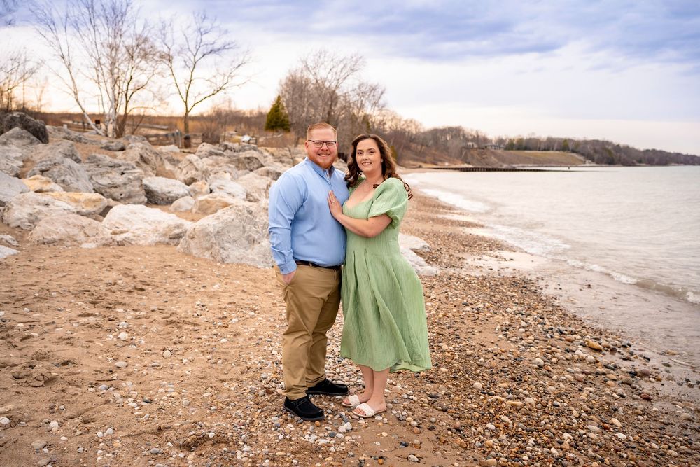 A couple poses together on a rocky beach shoreline with a wintry landscape behind them on a cloudy day.