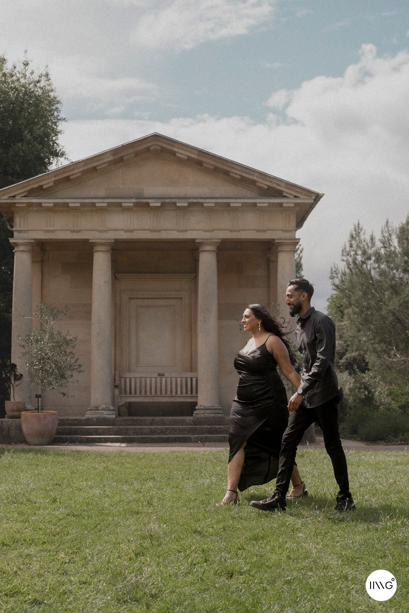 A couple strolls on a grassy lawn in front of a neoclassical building with columns and a triangular pediment.