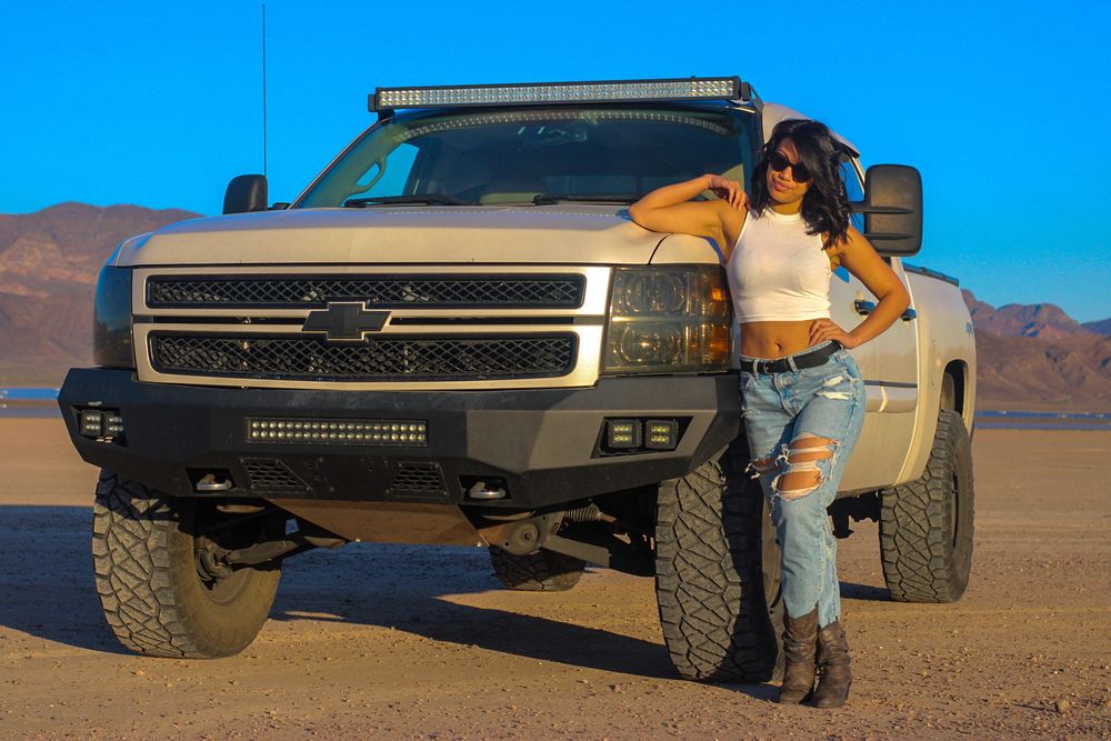 Woman in ripped jeans posing beside lifted Chevrolet Silverado truck in arid desert landscape.