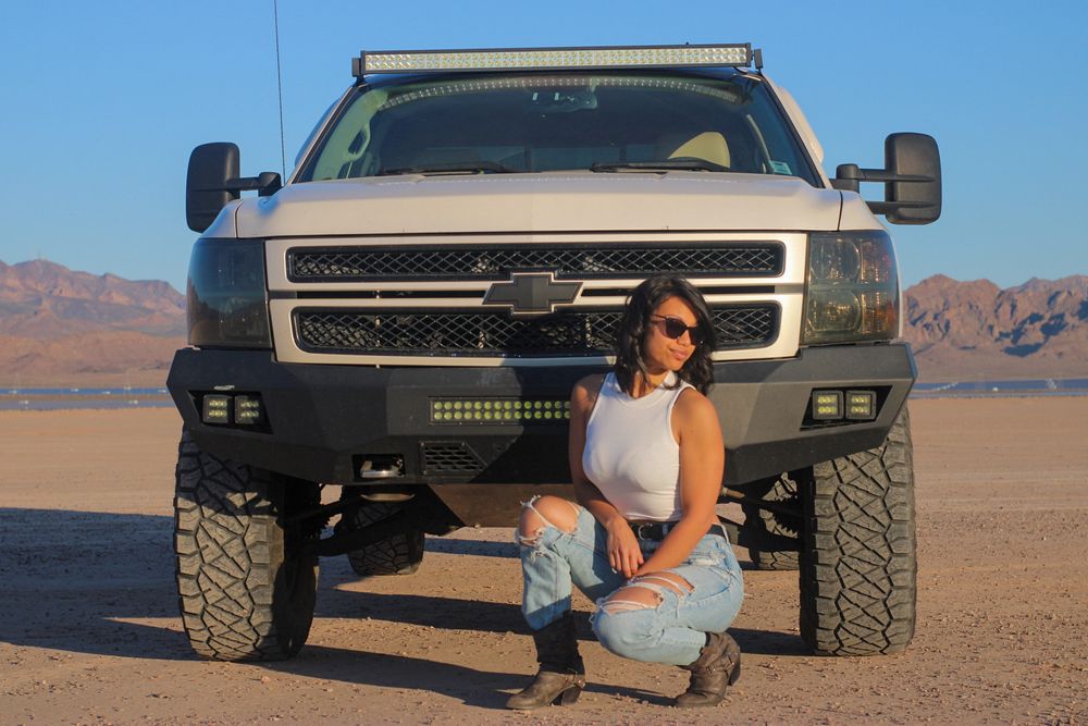 Woman crouching in front of lifted white Chevrolet truck on sandy desert terrain with mountains.