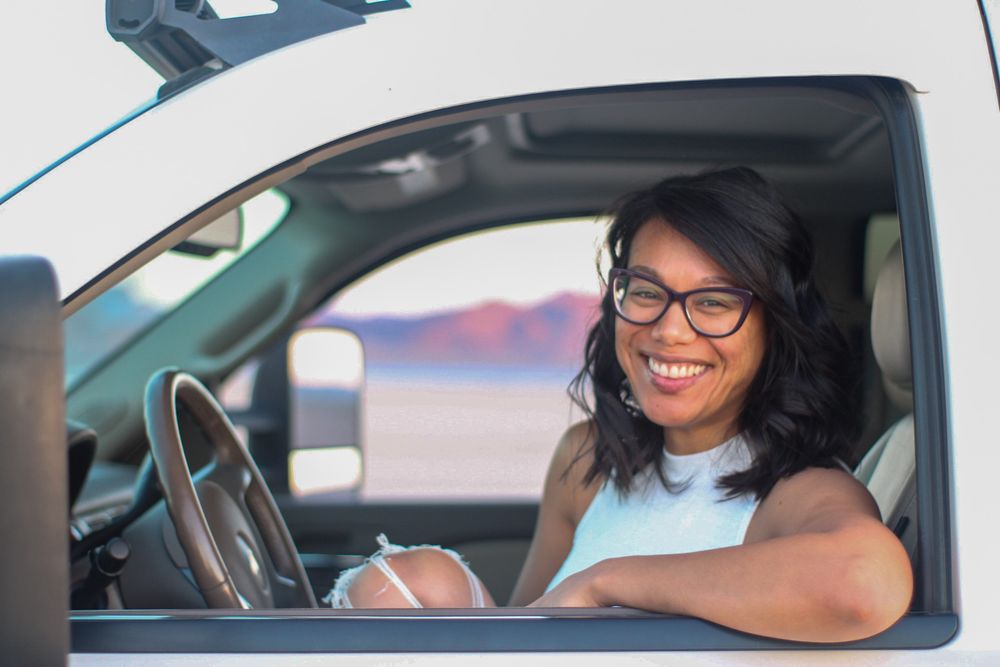 Smiling woman with glasses sitting in drivers seat of white pickup truck with desert landscape behind.