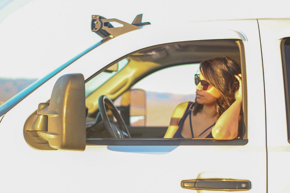 Woman wearing sunglasses sitting in drivers seat of white pickup truck viewed through open window.