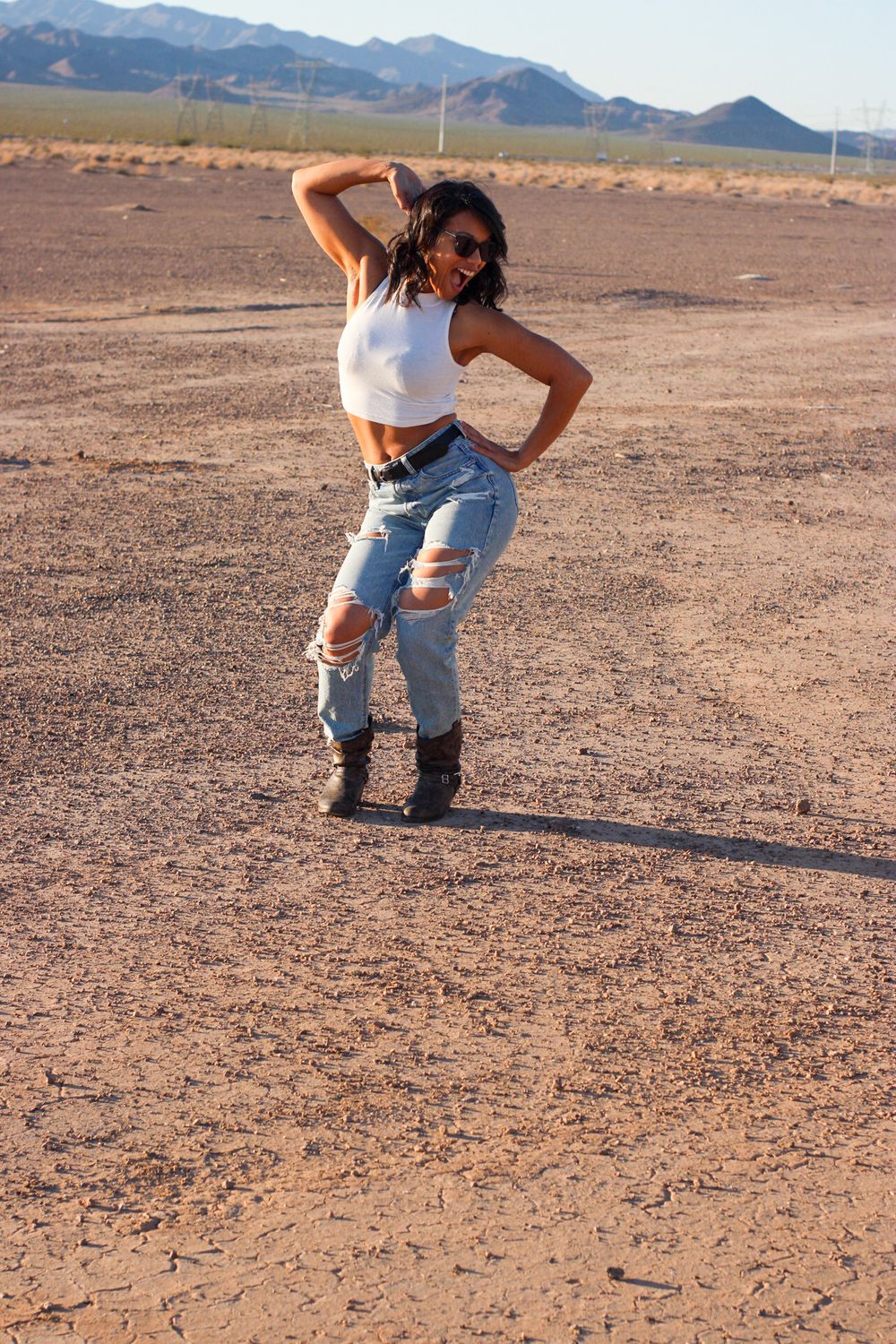 Woman in white crop top and ripped jeans posing in sunny desert landscape with mountains.