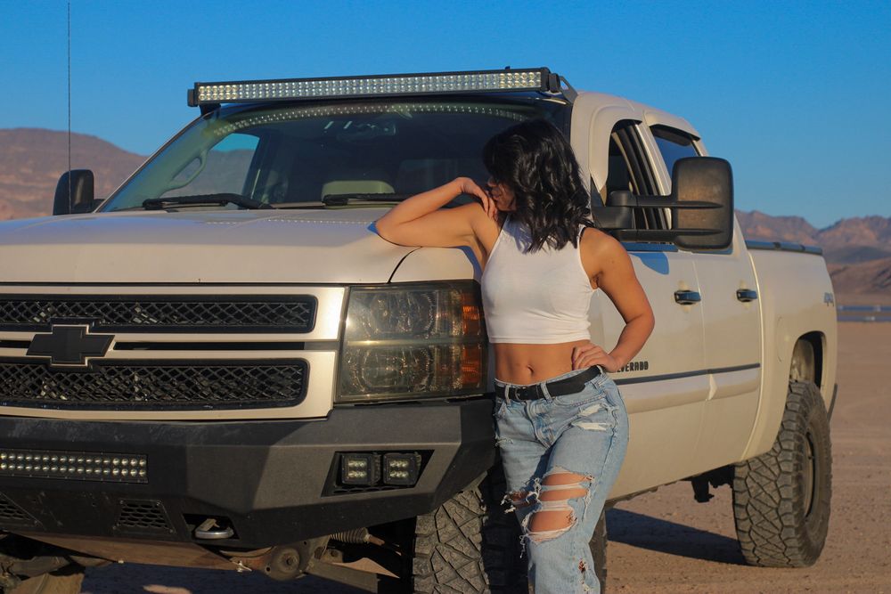 Woman in white crop top and ripped jeans posing beside lifted Chevy Silverado in desert.